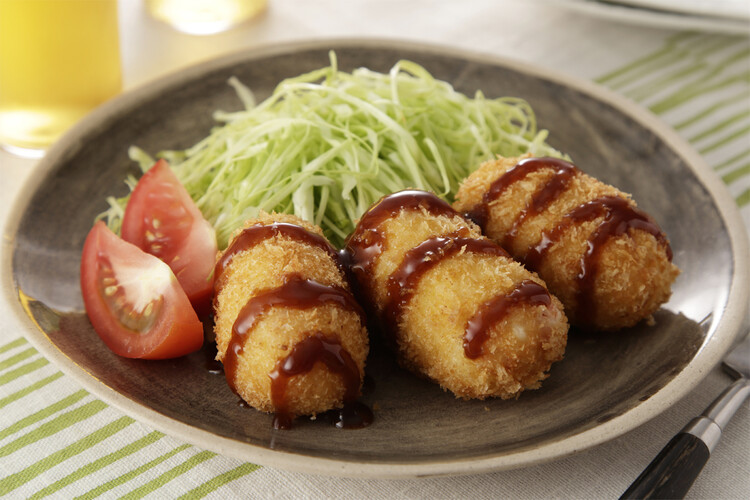 3 fried golden korokke (potato croquettes) drizzled in a tonkatsu glaze, sitting next to a small bed of shredded cabbage and two quarters of a tomato.